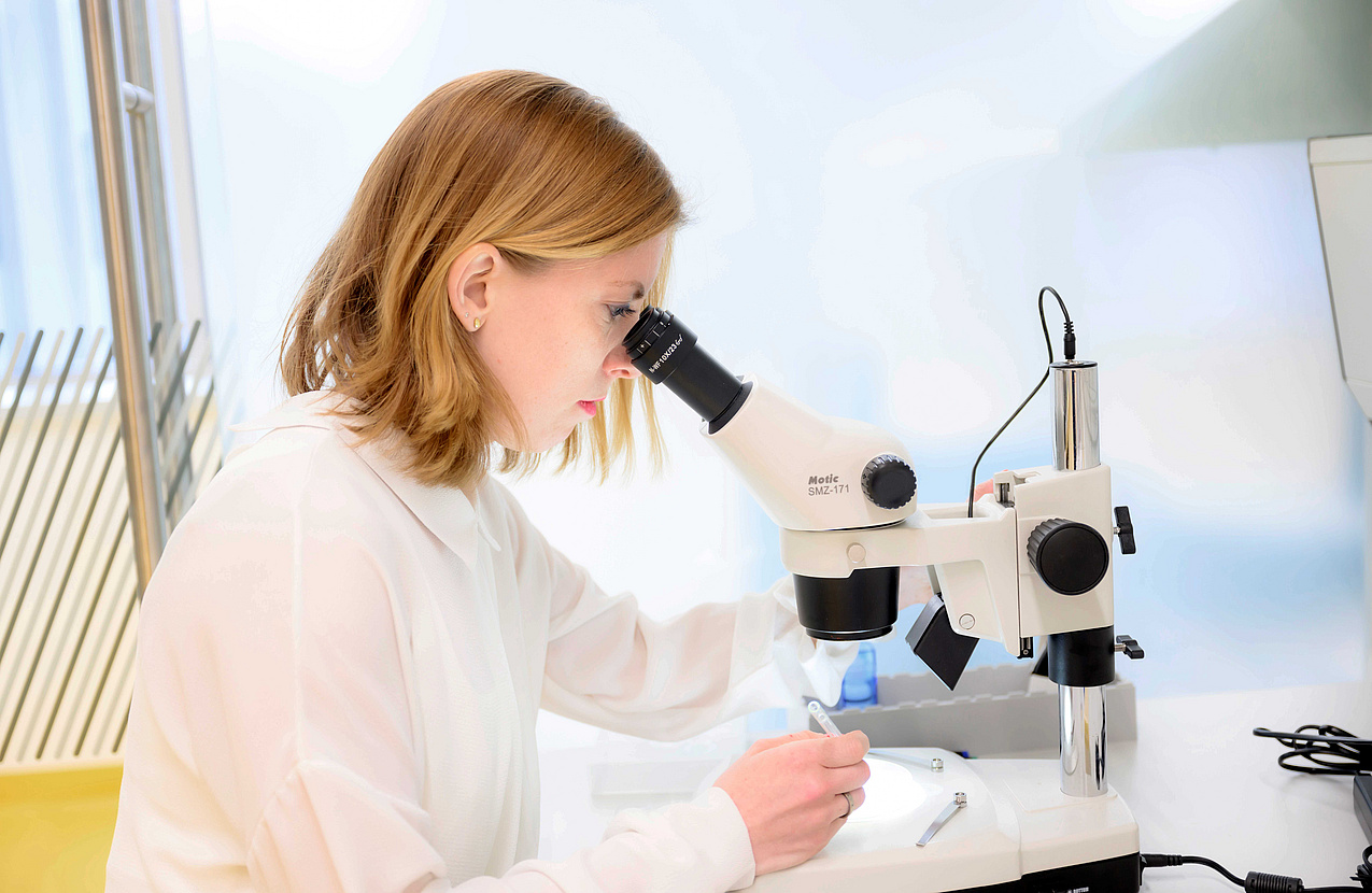 Eine Forscherin sitzt in einem Labor und begutachtet ein Objekt im Mikroskop.