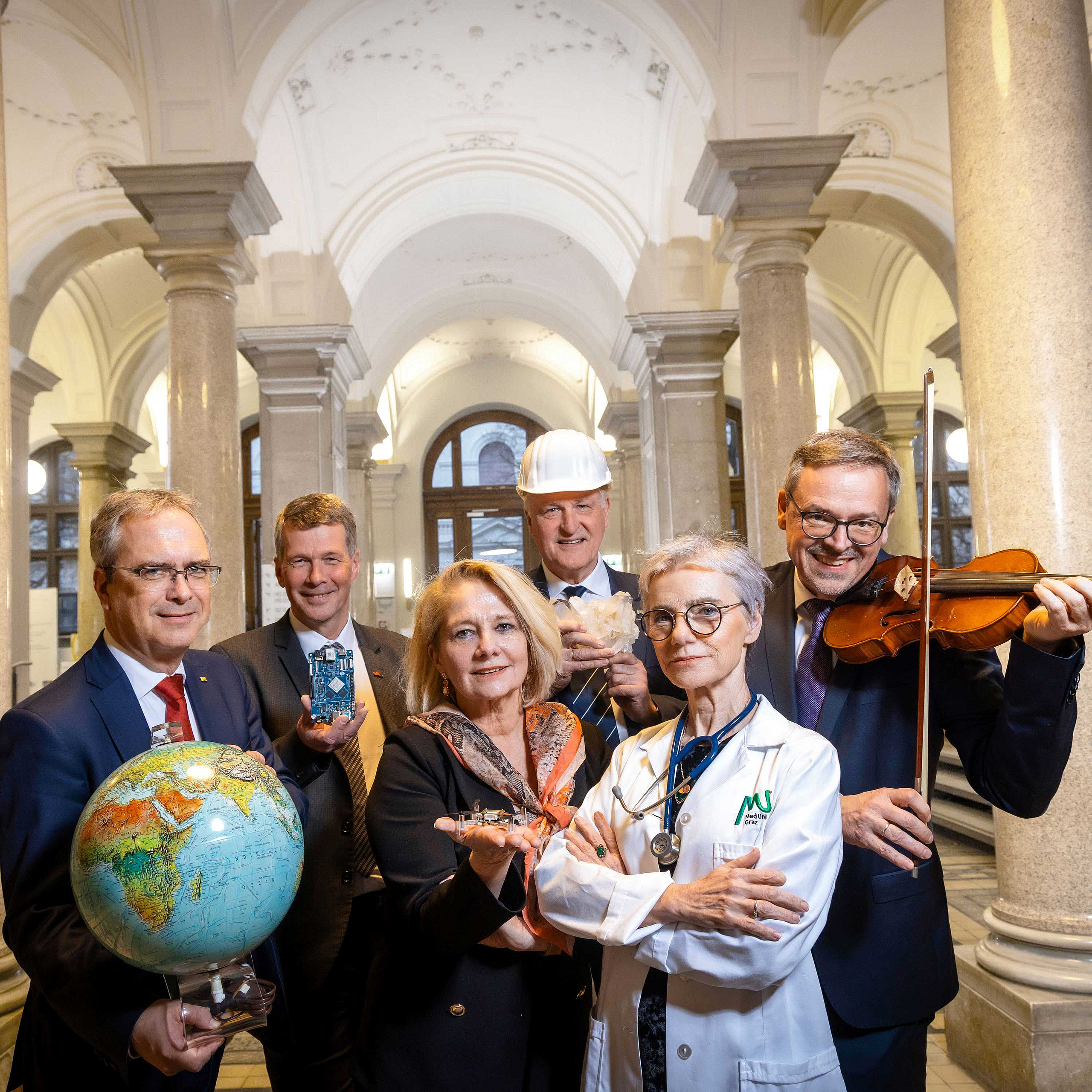 Die Rektor*innen stehen in der Universität Graz und halten Objekte, welche die Disziplinen ihrer Universität repräsentieren.