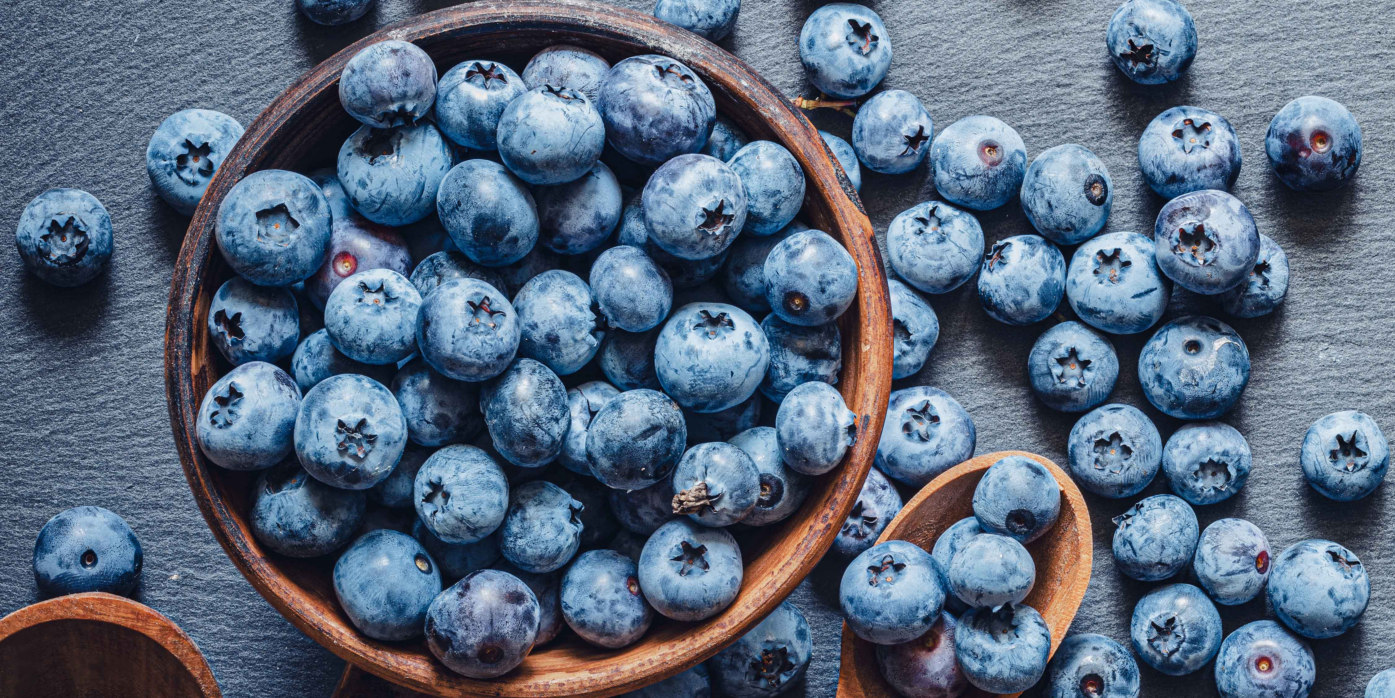 Viele Heidelbeeren auf blau-grauem Hintergrund, in einem Holzbecher und auf Holzlöffeln.