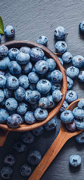 Viele Heidelbeeren auf blau-grauem Hintergrund, in einem Holzbecher und auf Holzlöffeln.